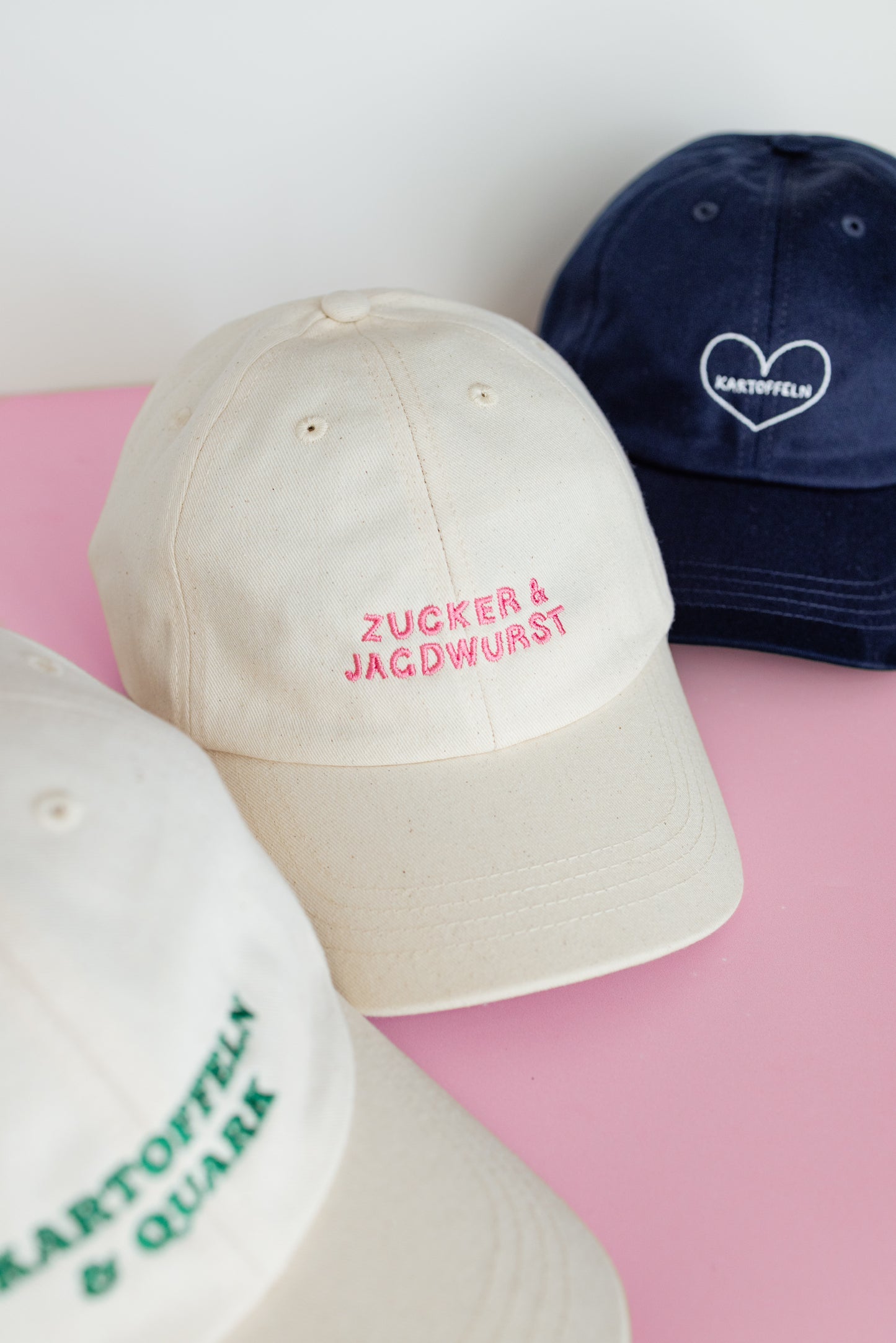 Drei Kappen: weiße Kappe mit pinker Stickerei ‚Zucker & Jagdwurst‘, weiße Kappe mit pinker Stickerei ‚Zucker & Jagdwurst‘, dunkelblaue Kappe mit weißem Herz und Text ‚Kartoffeln‘